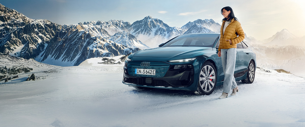 Eine Frau steht neben einem grünen Audi auf verschneitem Bergplateau mit hohen, schneebedeckten Gipfeln im Hintergrund.