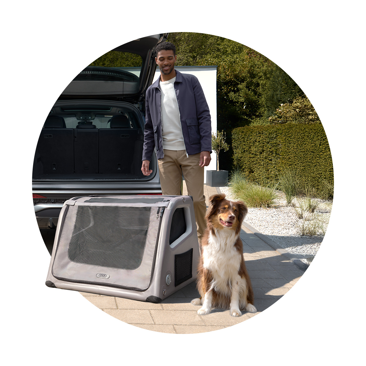 Ein Mann steht am offenen Kofferraum eines Audi, daneben eine Hundebox und ein Hund sitzt auf dem Gehweg.